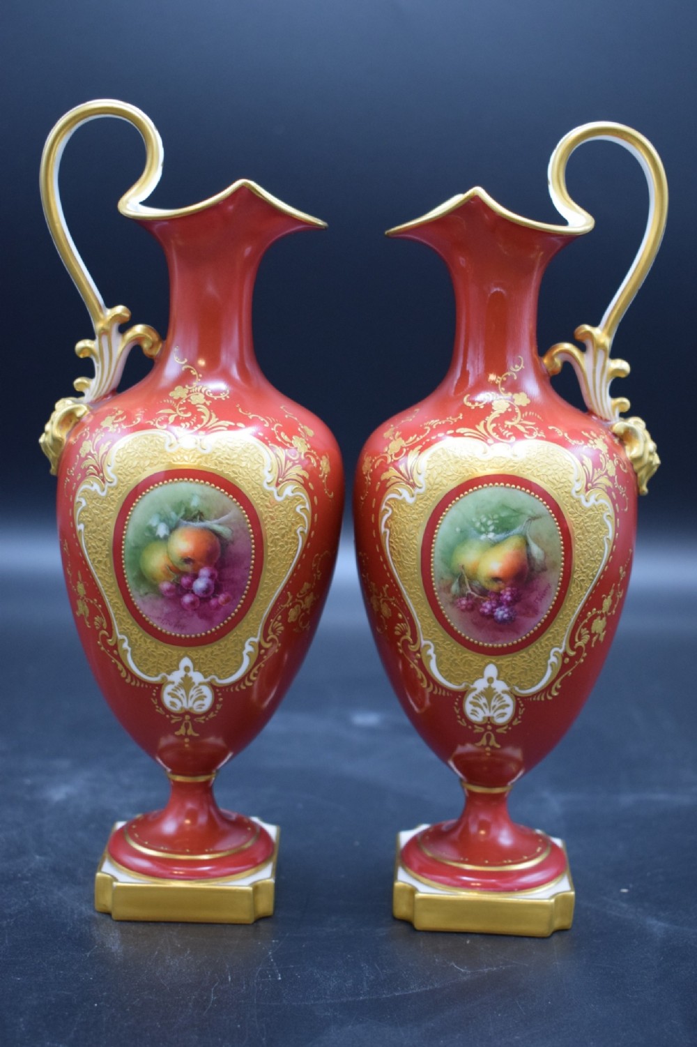 A pair of crimson red porcelain ewers featuring hand-painted fruit vignettes enclosed within gilt scrollwork borders and loop handles on square pedestal bases.