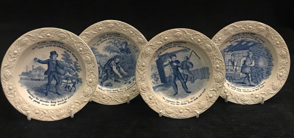 A set of four circular cream pearlware nursery plates featuring blue transfer-printed scenes of historical trades enclosed by moulded floral borders.