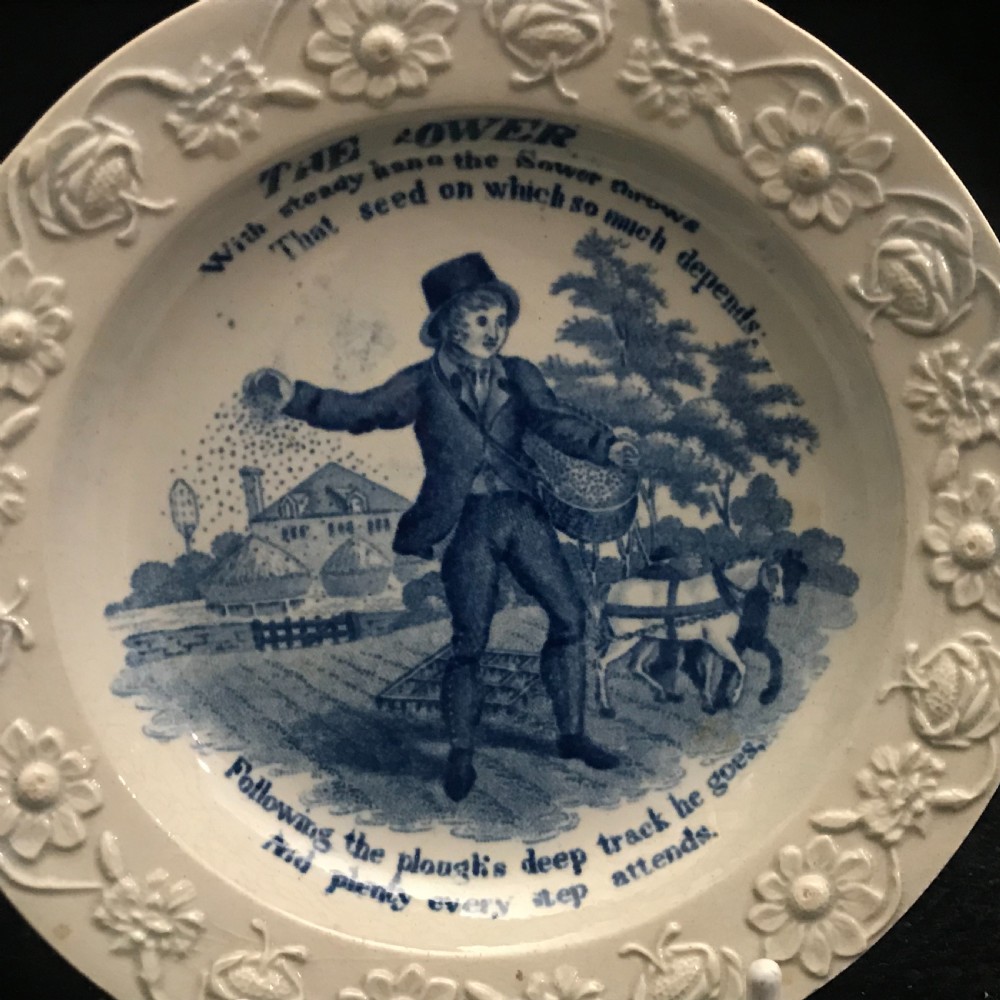 A set of four circular cream pearlware nursery plates featuring blue transfer-printed scenes of historical trades enclosed by moulded floral borders. View II