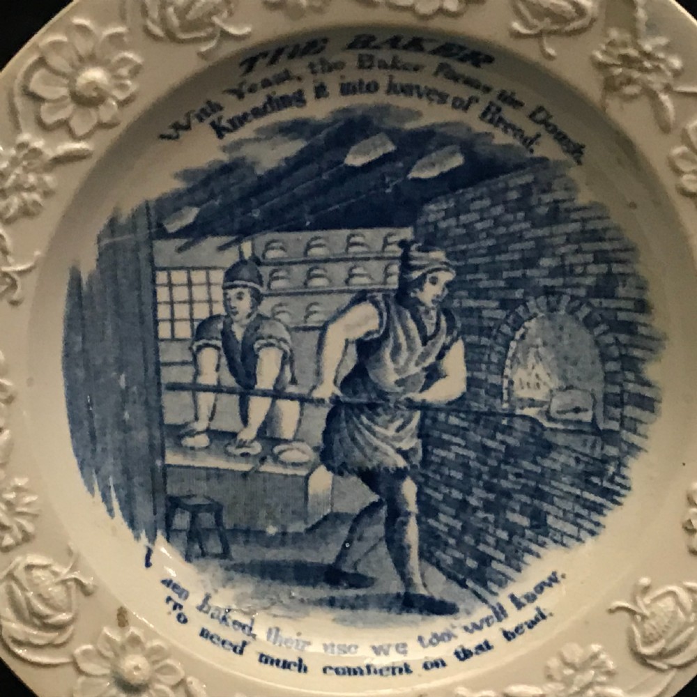 A set of four circular cream pearlware nursery plates featuring blue transfer-printed scenes of historical trades enclosed by moulded floral borders. View V