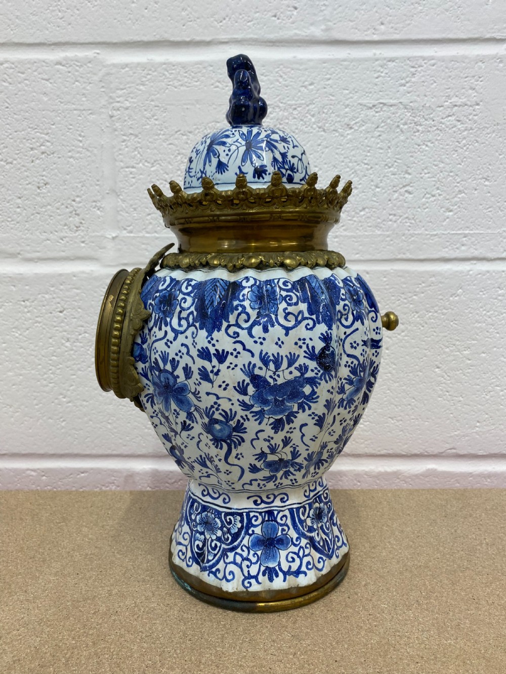 A blue and white floral patterned ceramic vase clock with a fitted lid, featuring an integrated brass clock face with Roman numerals. View II