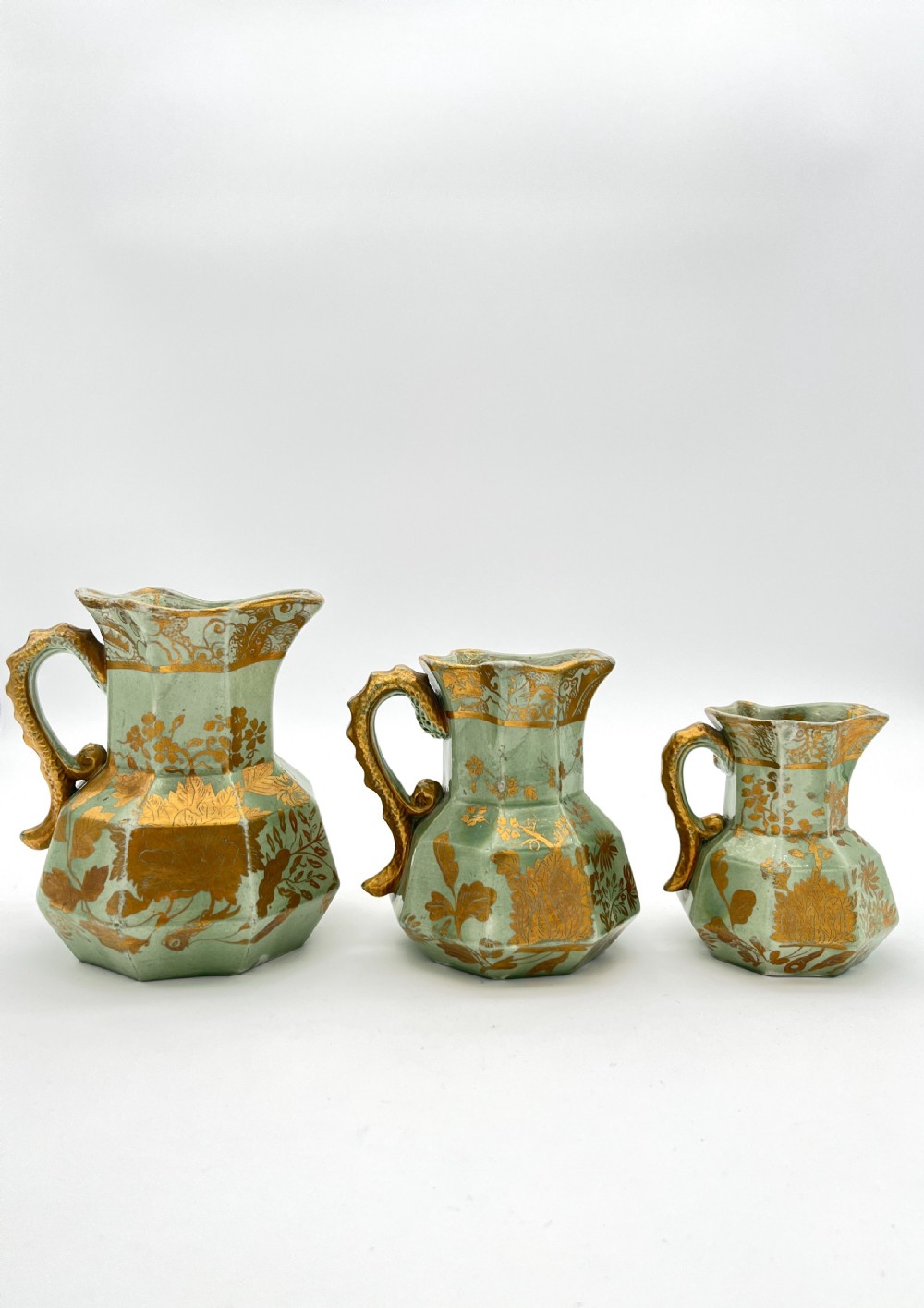 A set of three graduated octagonal sage green ironstone jugs decorated with gilded floral motifs and ornate moulded scroll handles.