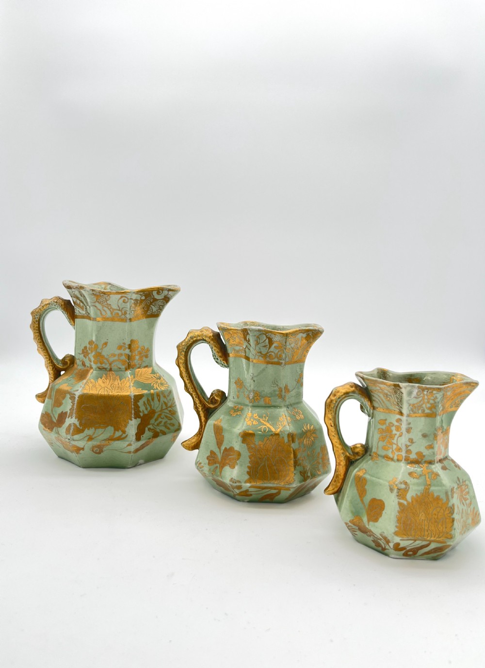 A set of three graduated octagonal sage green ironstone jugs decorated with gilded floral motifs and ornate moulded scroll handles. View II