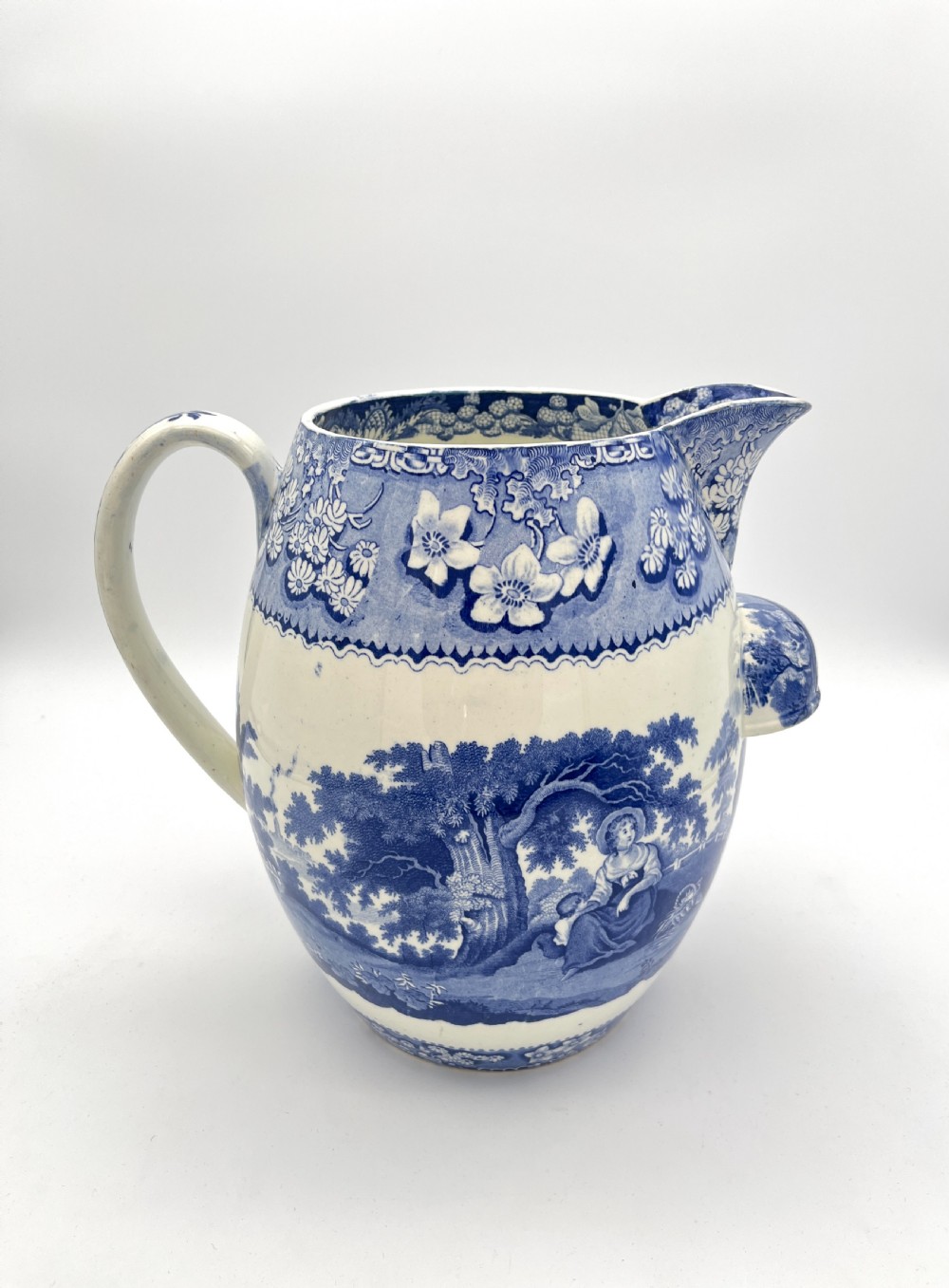 An antique blue and white earthenware footbath jug decorated with a floral upper band and a printed pastoral landscape scene.