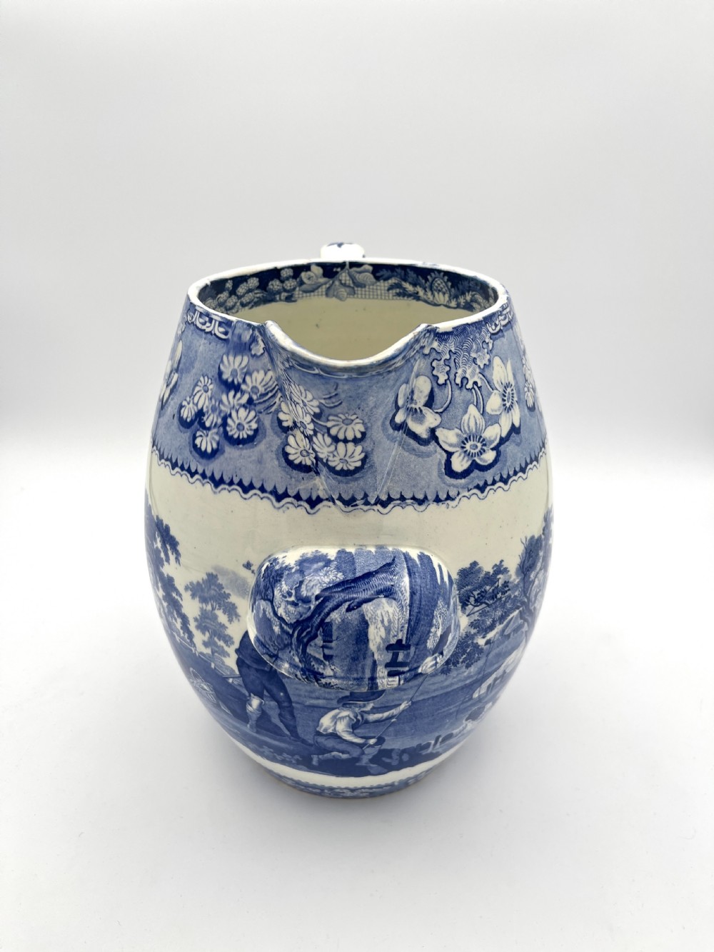 An antique blue and white earthenware footbath jug decorated with a floral upper band and a printed pastoral landscape scene. View II