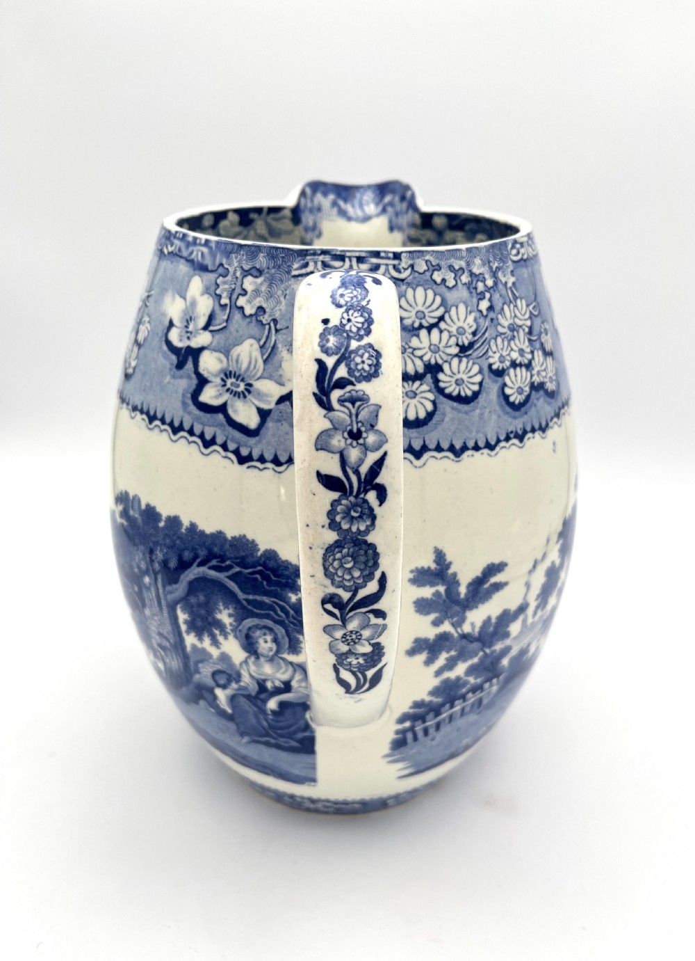 An antique blue and white earthenware footbath jug decorated with a floral upper band and a printed pastoral landscape scene. View VI