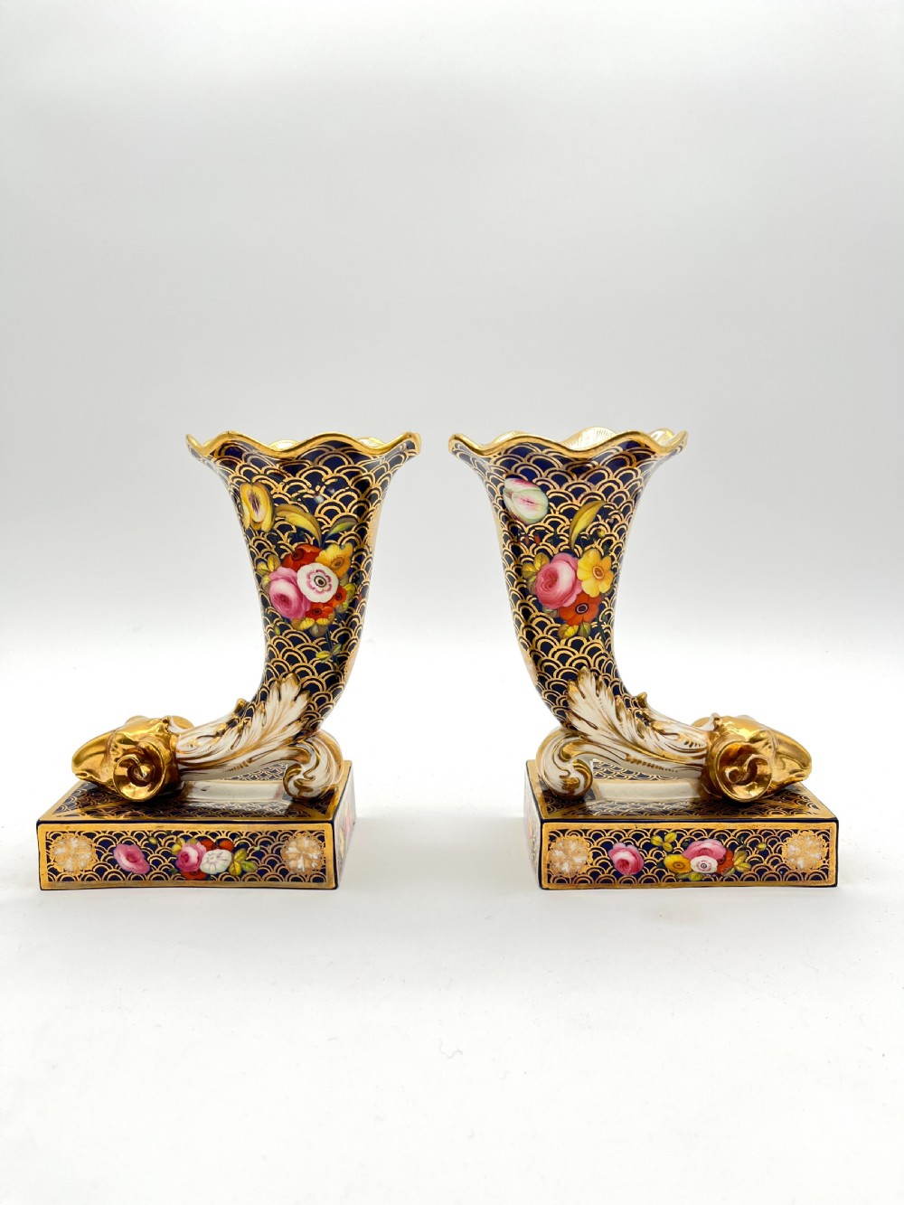 A pair of Regency porcelain cornucopia vases featuring deep cobalt blue fishscale patterning, gold leaf trim, and hand-painted floral panels on scroll bases.