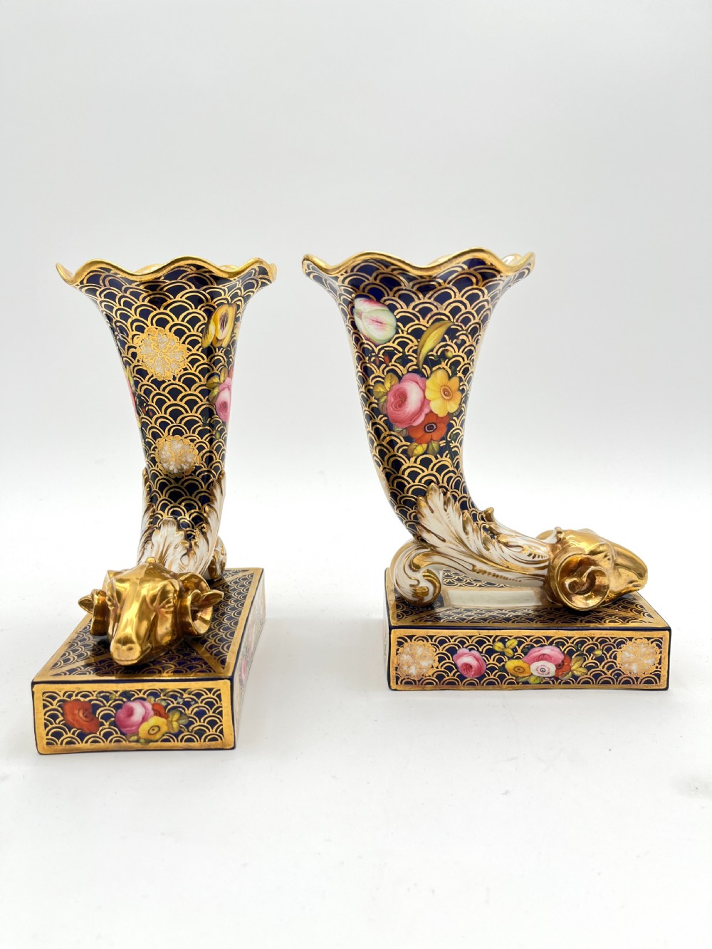 A pair of Regency porcelain cornucopia vases featuring deep cobalt blue fishscale patterning, gold leaf trim, and hand-painted floral panels on scroll bases. View VI