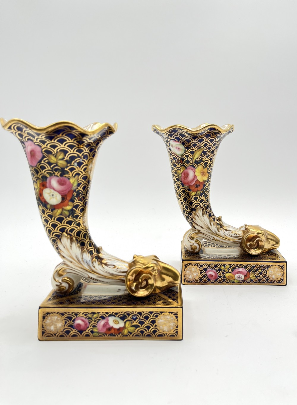 A pair of Regency porcelain cornucopia vases featuring deep cobalt blue fishscale patterning, gold leaf trim, and hand-painted floral panels on scroll bases. View VII