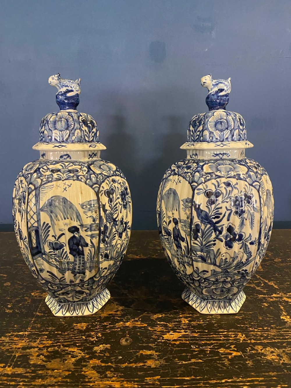 A pair of octagonal blue and white tin-glazed earthenware lidded vases decorated with hand-painted chinoiserie scenes and animal finials.