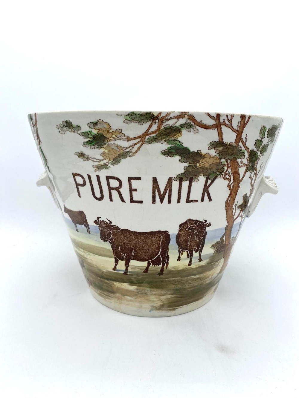 A cream earthenware Victorian milk pail decorated with brown painted cows under trees and lettering stating Pure Milk. View II