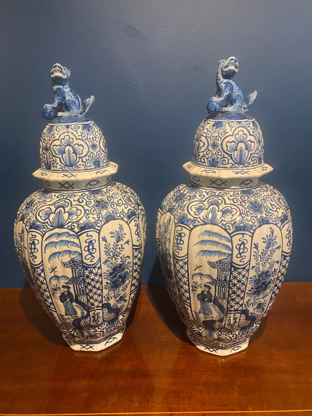 A pair of mid-19th century ribbed blue and white tin-glazed earthenware lidded vases hand-painted with oriental landscapes and topped with seated lion finials.