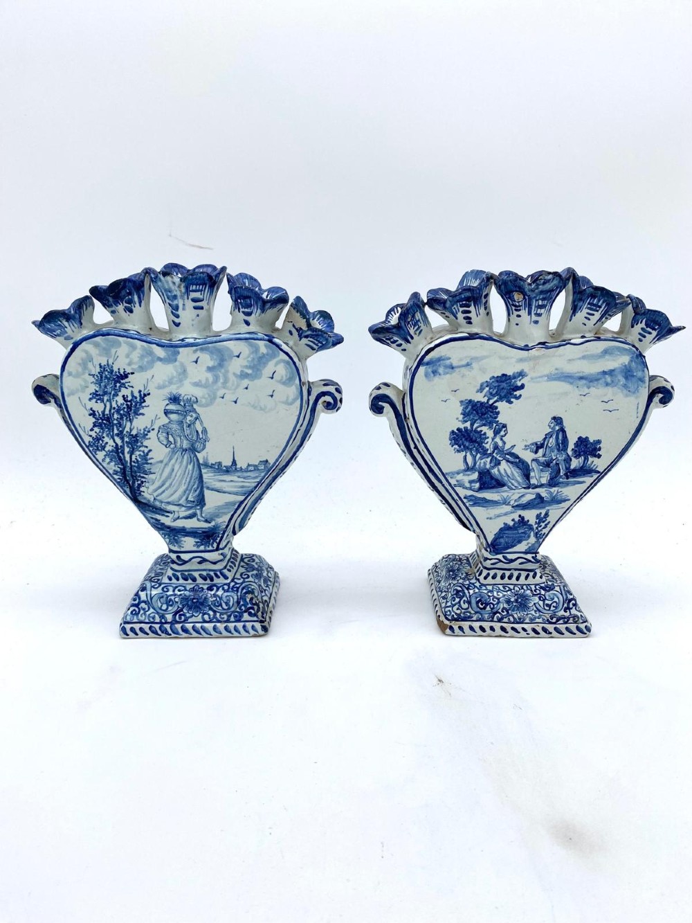 A pair of blue and white heart-shaped ceramic tulipieres featuring multiple spouts and hand-painted Dutch figures in landscape settings.