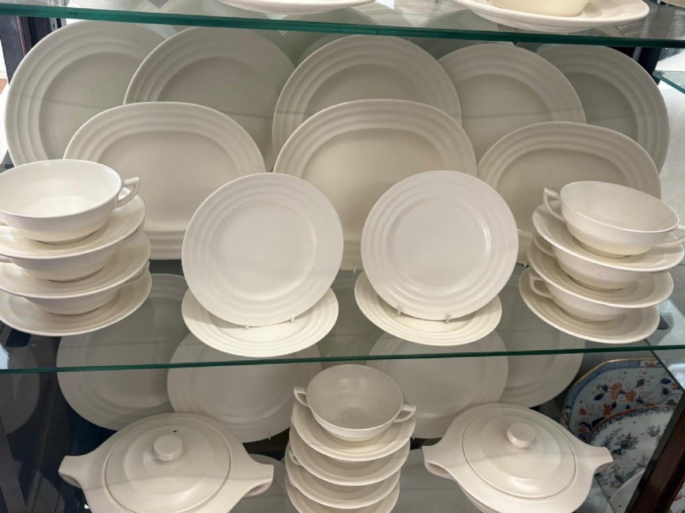 A comprehensive Art Deco off-white ceramic dinner service by Keith Murray for Wedgwood, featuring tiered plates, platters, covered tureens, and soup bowls with ribbed concentric detailing. View V