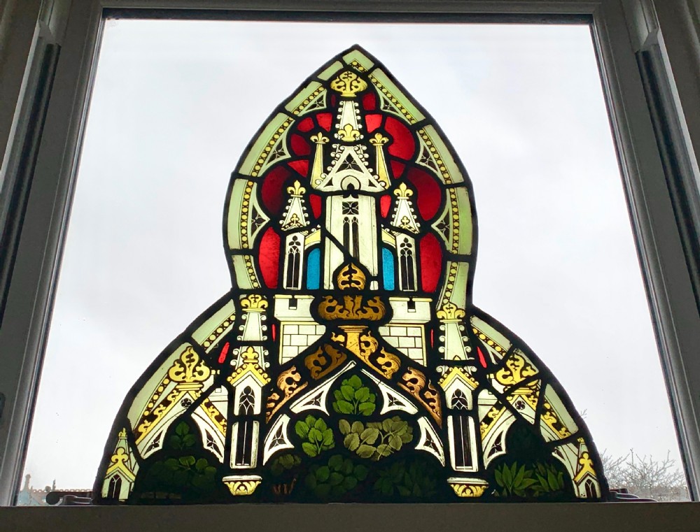 An arched Gothic Revival stained glass panel featuring painted architectural spires, foliate patterns, and lead cames in red, green, yellow, and clear glass.