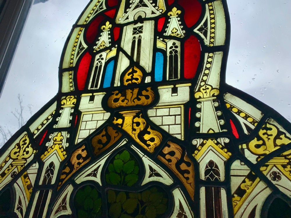 An arched Gothic Revival stained glass panel featuring painted architectural spires, foliate patterns, and lead cames in red, green, yellow, and clear glass. View V