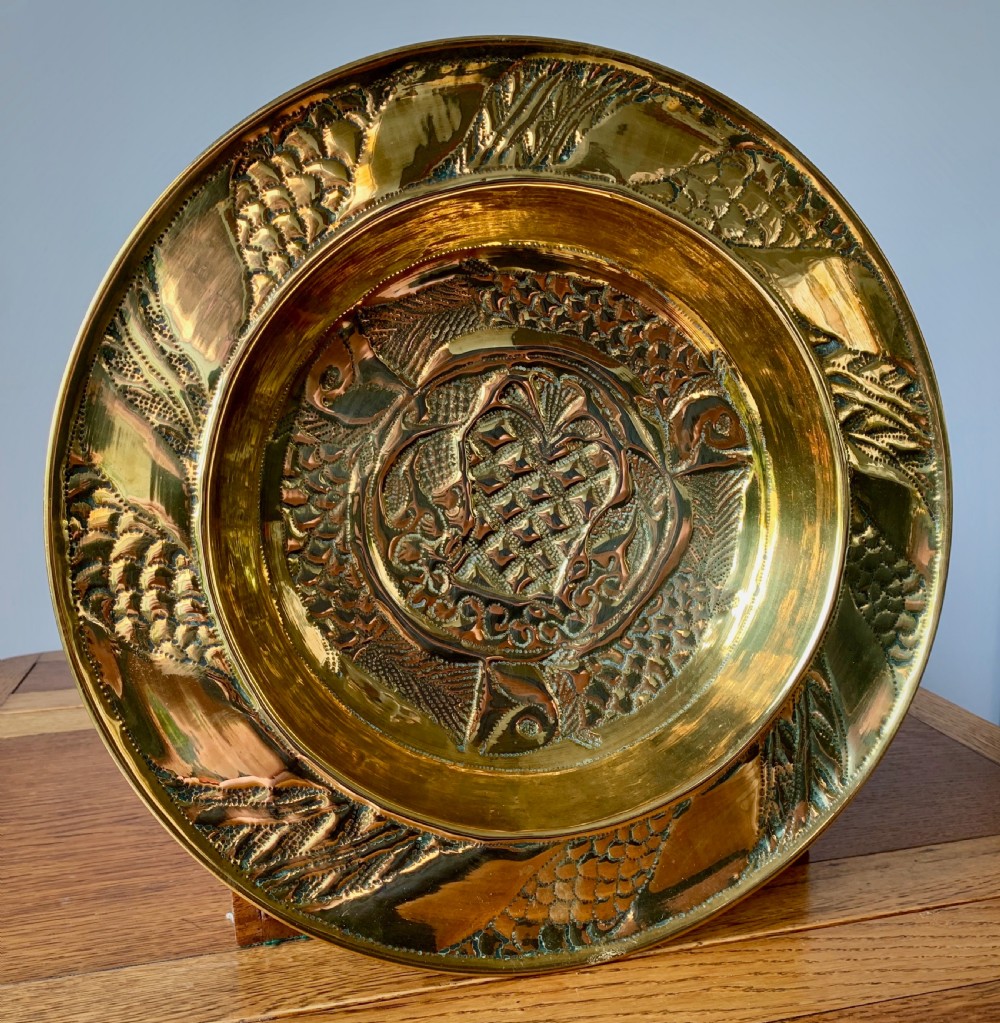 A circular Arts and Crafts hand-hammered brass charger featuring repousse decoration of three fish surrounding a central fountain design.