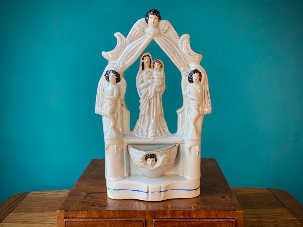 An antique Staffordshire earthenware holy water stoup with figures of the Madonna and Child under an arch flanked by angels in cream glaze with blue and brown painted details.