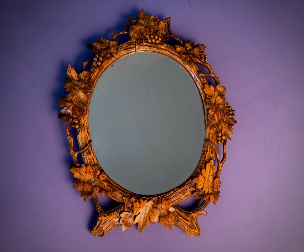 An early 20th-century oval wall mirror featuring a carved wooden frame decorated with grapevines, leaves, and branch motifs around a bevelled glass mirror plate.