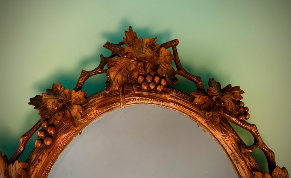 An early 20th-century oval wall mirror featuring a carved wooden frame decorated with grapevines, leaves, and branch motifs around a bevelled glass mirror plate. View II