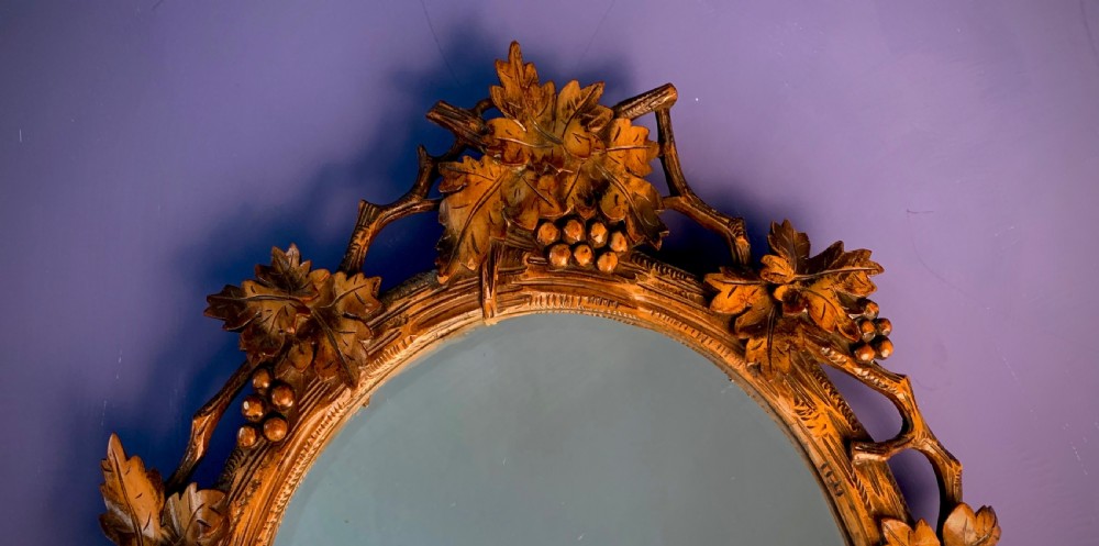 An early 20th-century oval wall mirror featuring a carved wooden frame decorated with grapevines, leaves, and branch motifs around a bevelled glass mirror plate. View IV