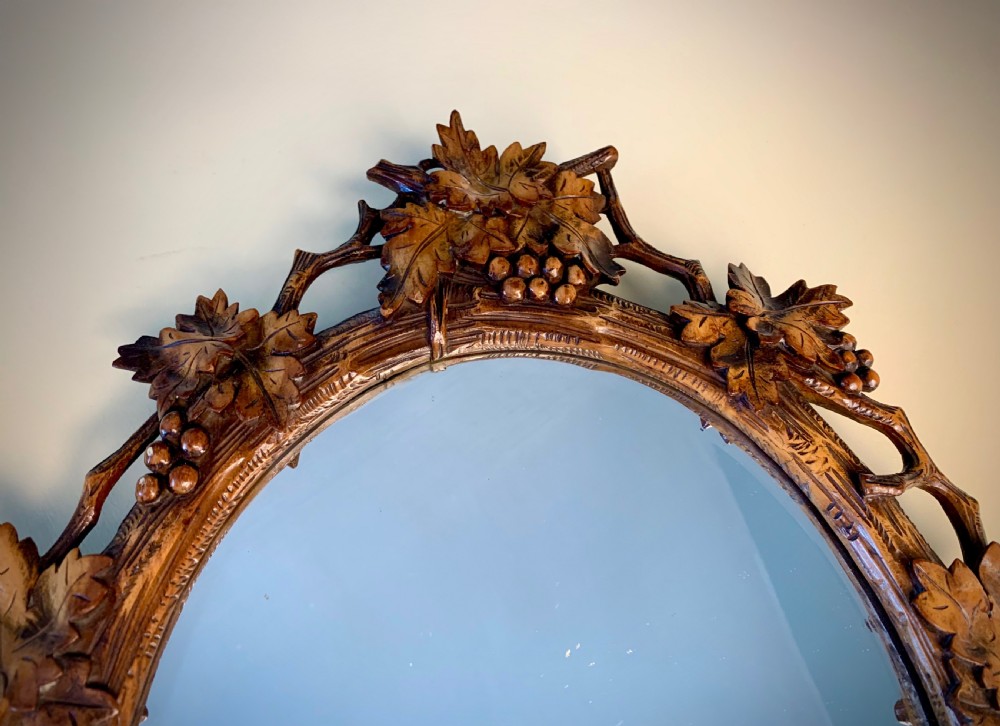 An early 20th-century oval wall mirror featuring a carved wooden frame decorated with grapevines, leaves, and branch motifs around a bevelled glass mirror plate. View V