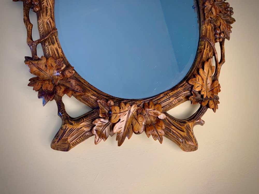 An early 20th-century oval wall mirror featuring a carved wooden frame decorated with grapevines, leaves, and branch motifs around a bevelled glass mirror plate. View X