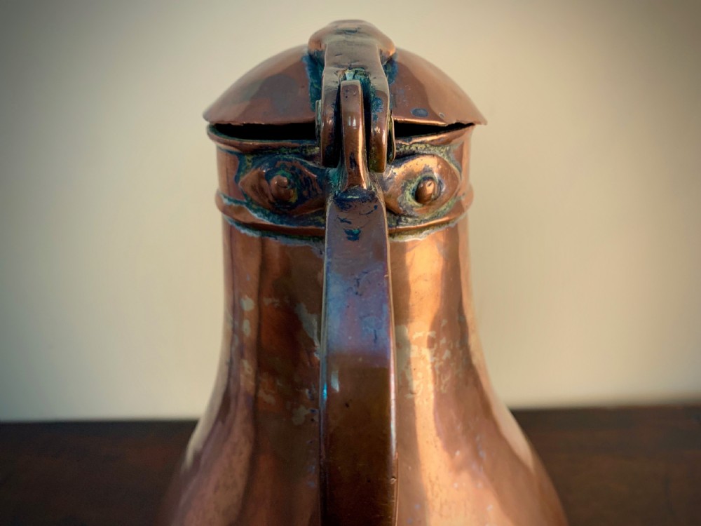 A large hand-hammered copper pitcher featuring a tapered neck, rounded body, strap handle, and a hinged lid. View V