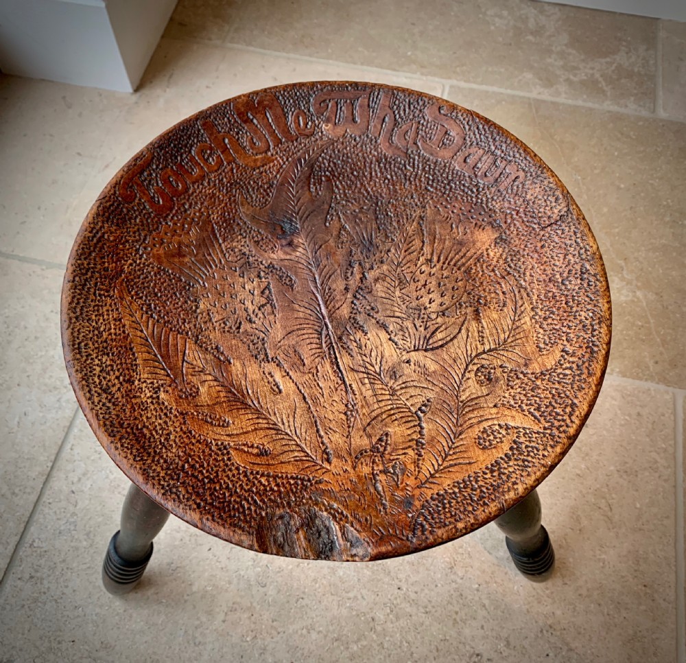 A 19th-century Scottish tripod stool crafted from dark brown wood with a circular seat featuring burnt pokerwork text and a Scottish thistle motif. View VI