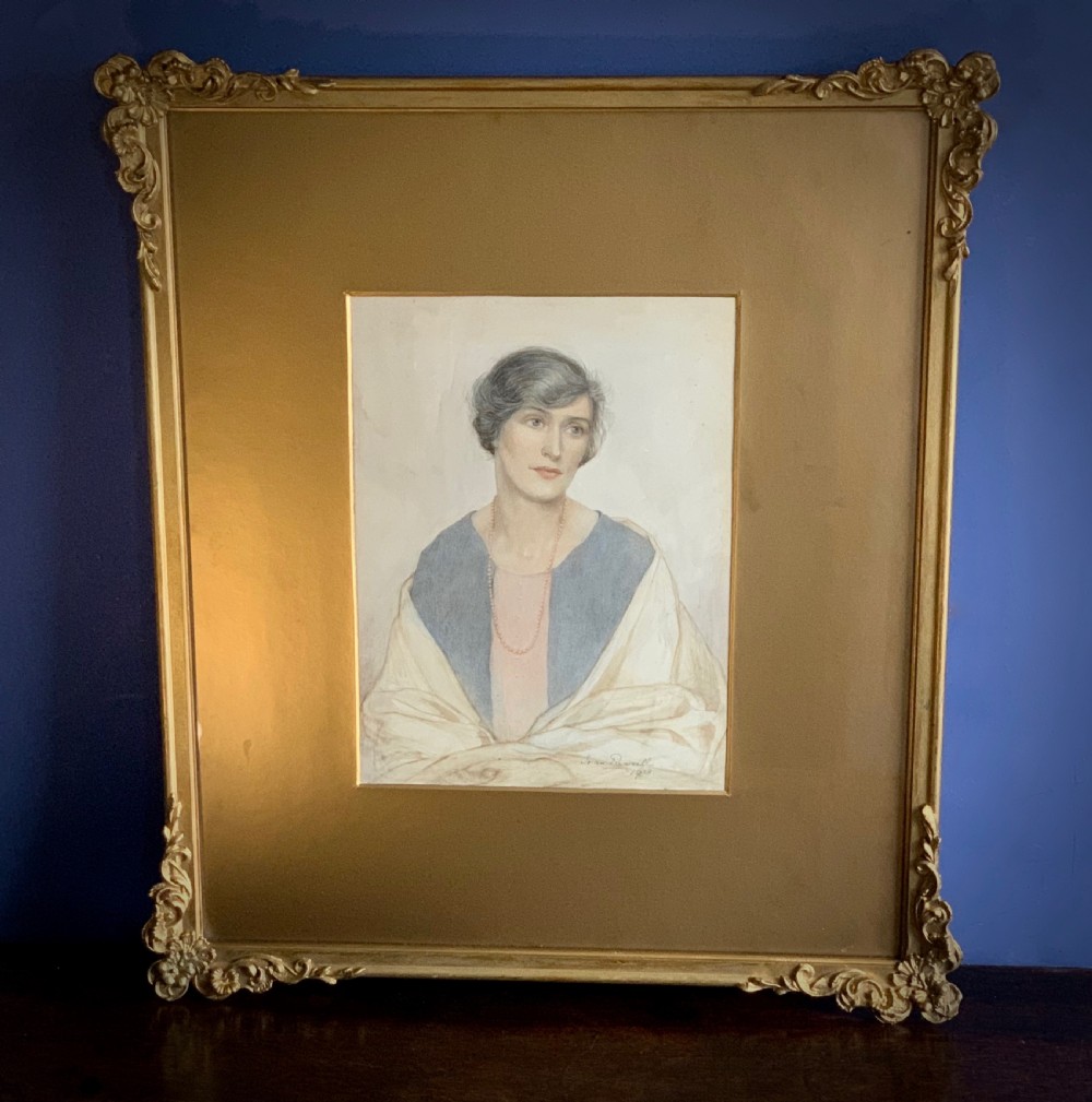 A framed 1928 watercolour painting of a young woman with short dark hair, wearing a blue jacket and shawl, set within a wide gold mount and ornate gilded frame.