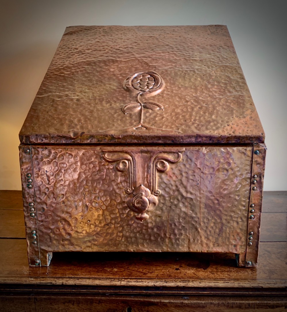A hand-hammered copper log box featuring an embossed stylised floral motif on the sloping lid and front panel, secured with riveted corner straps.