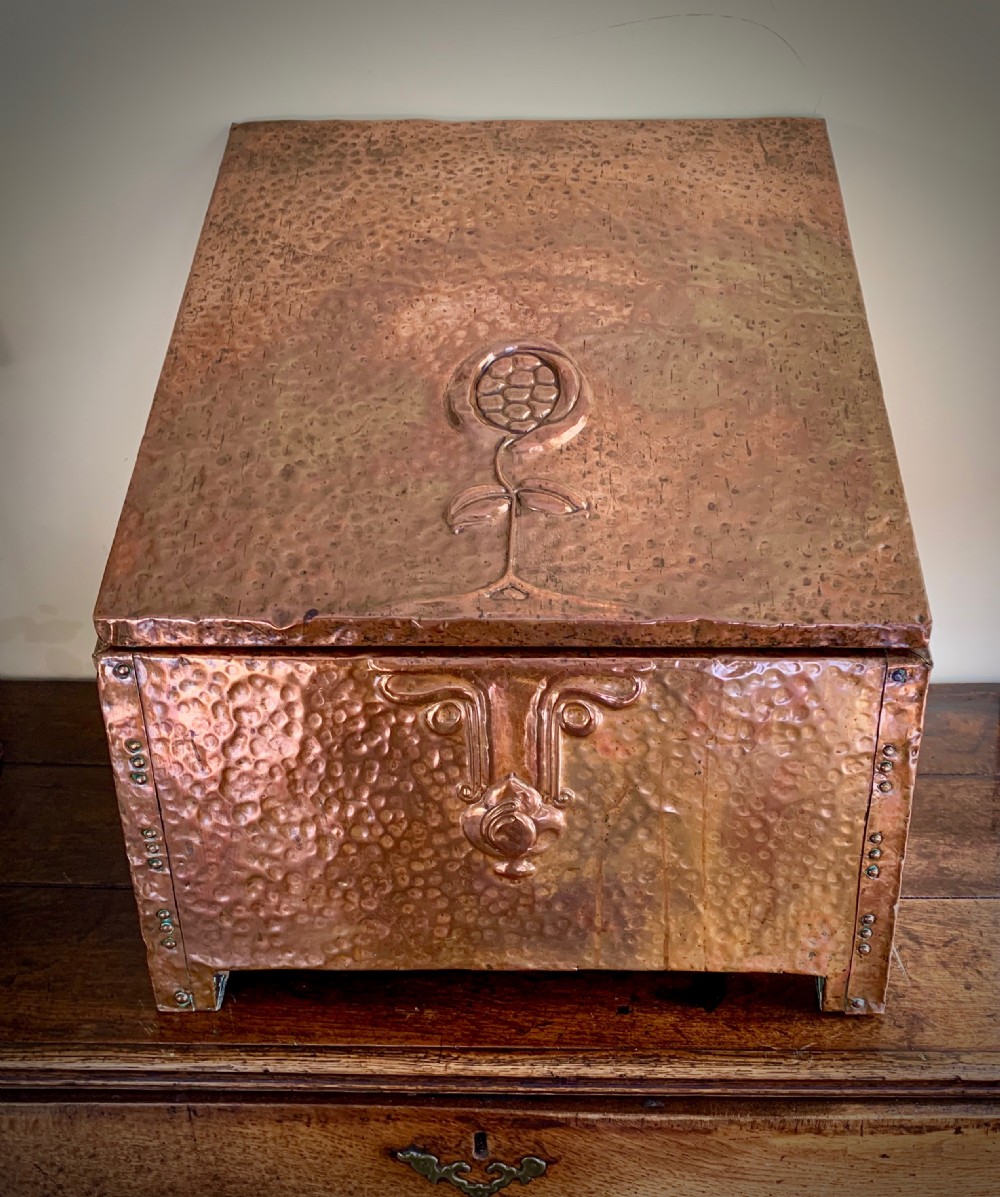 A hand-hammered copper log box featuring an embossed stylised floral motif on the sloping lid and front panel, secured with riveted corner straps. View II
