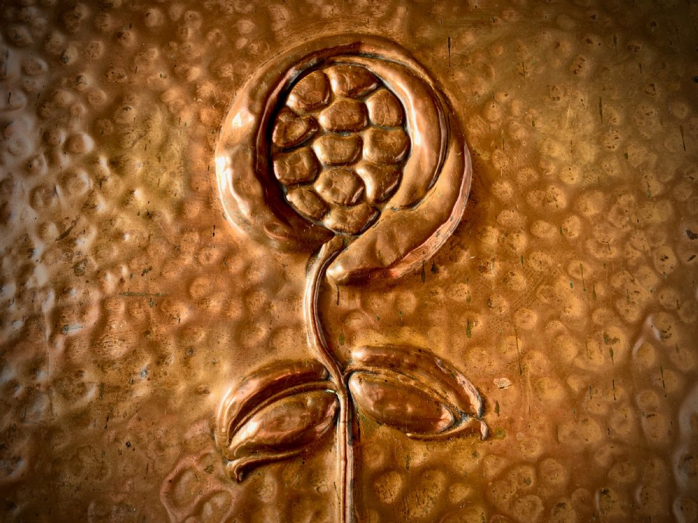 A hand-hammered copper log box featuring an embossed stylised floral motif on the sloping lid and front panel, secured with riveted corner straps. View III
