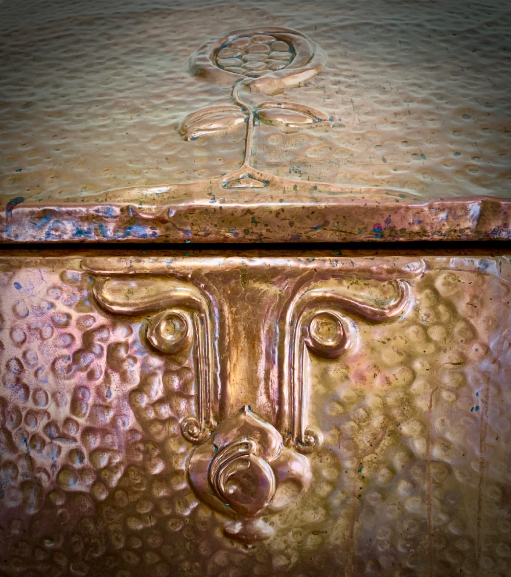 A hand-hammered copper log box featuring an embossed stylised floral motif on the sloping lid and front panel, secured with riveted corner straps. View IV