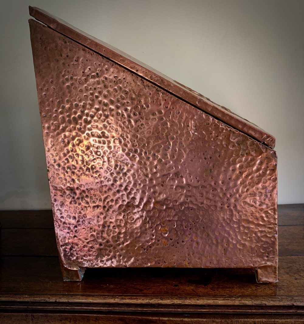 A hand-hammered copper log box featuring an embossed stylised floral motif on the sloping lid and front panel, secured with riveted corner straps. View VI
