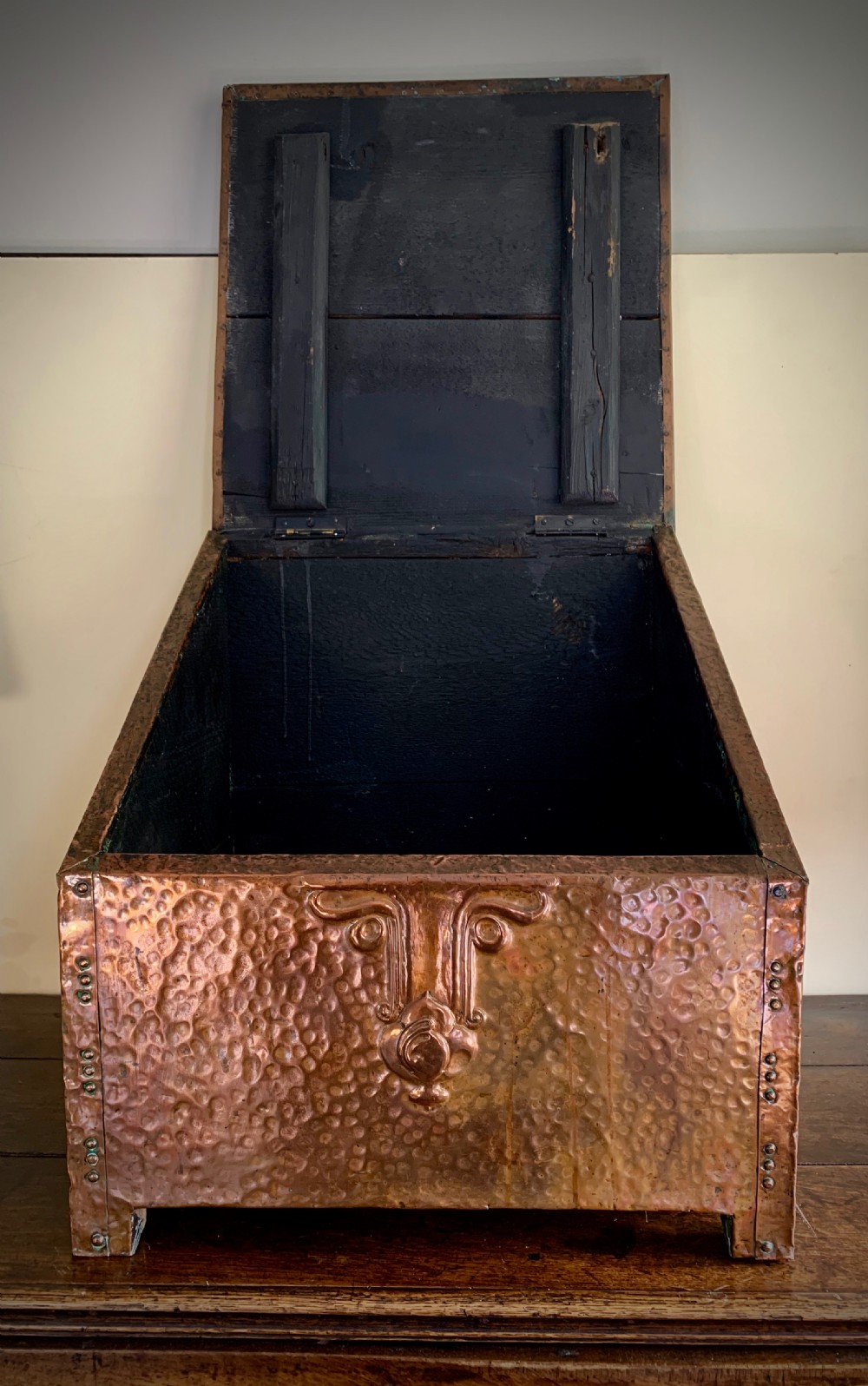 A hand-hammered copper log box featuring an embossed stylised floral motif on the sloping lid and front panel, secured with riveted corner straps. View IX