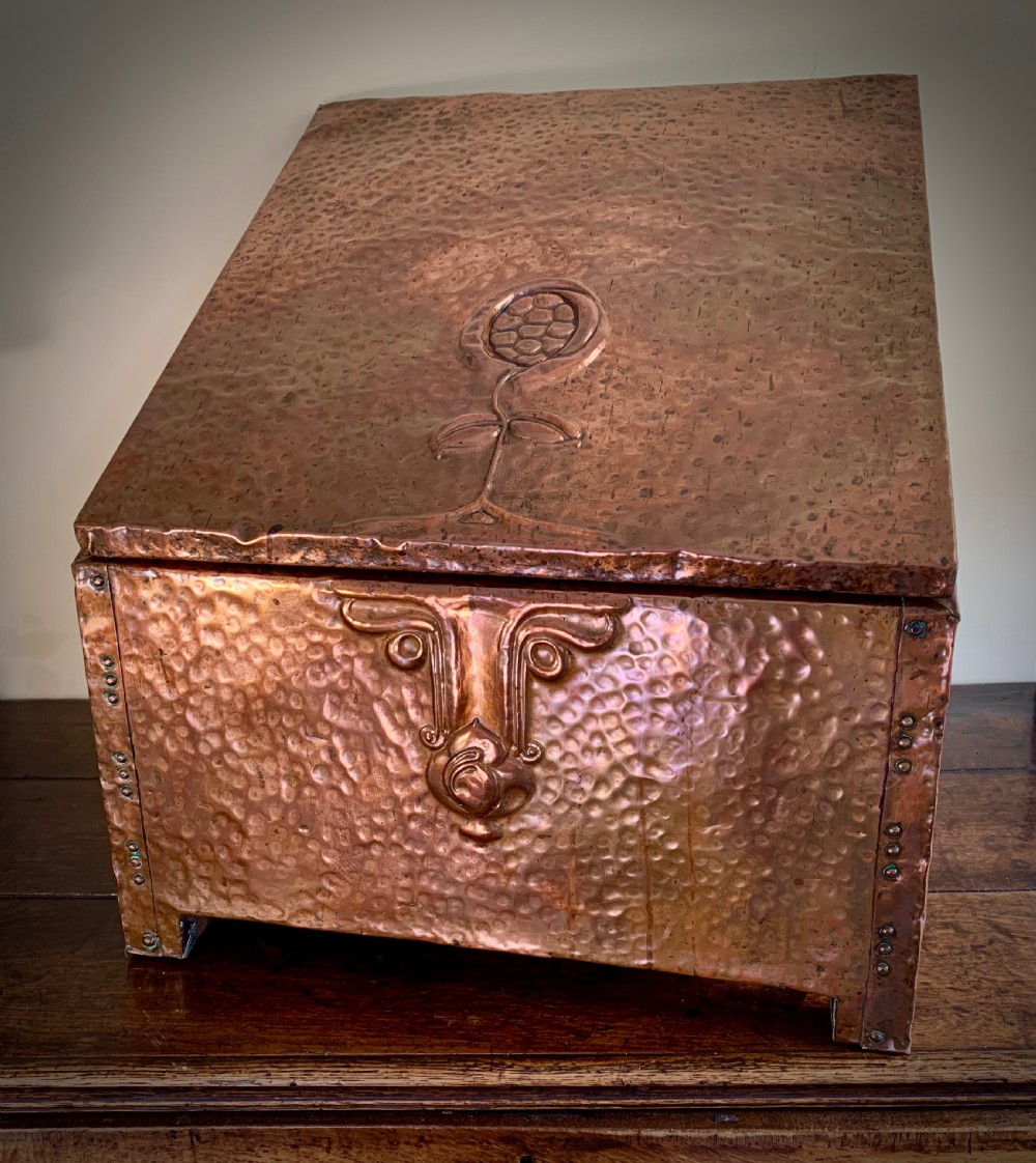 A hand-hammered copper log box featuring an embossed stylised floral motif on the sloping lid and front panel, secured with riveted corner straps. View X