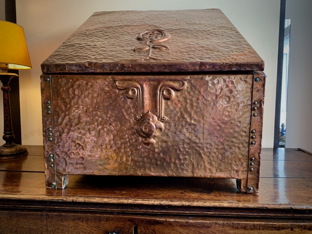 A hand-hammered copper log box featuring an embossed stylised floral motif on the sloping lid and front panel, secured with riveted corner straps. View XI