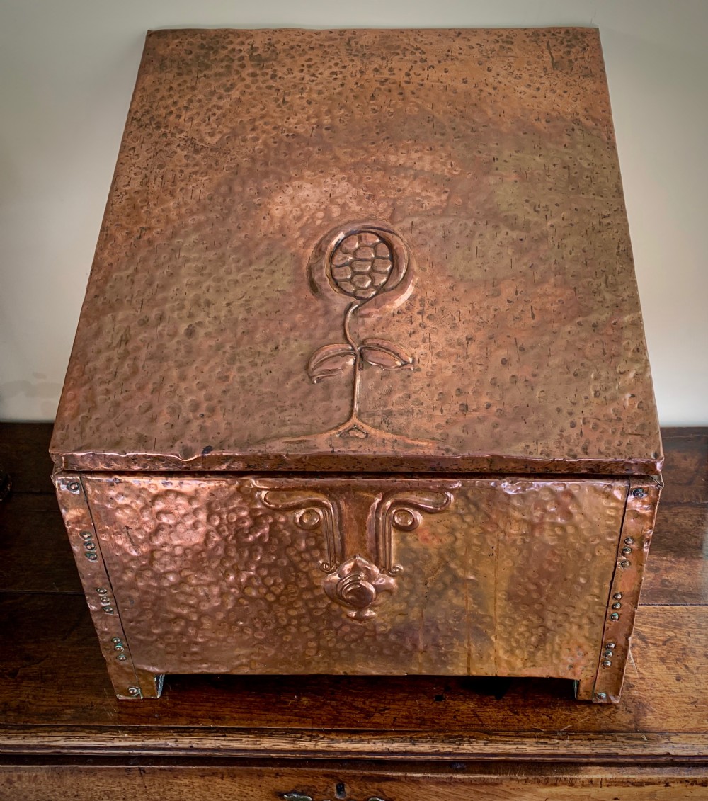 A hand-hammered copper log box featuring an embossed stylised floral motif on the sloping lid and front panel, secured with riveted corner straps. View XIII