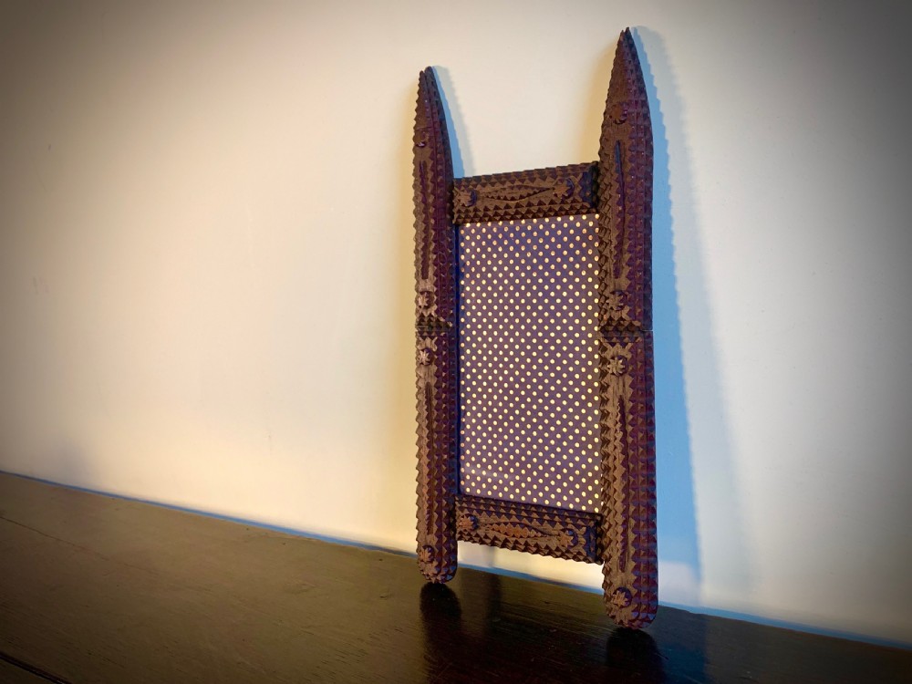 A dark brown hand-carved tramp art wooden picture frame featuring notch-carved geometric borders and pointed finials encasing a monochrome portrait photograph. View III