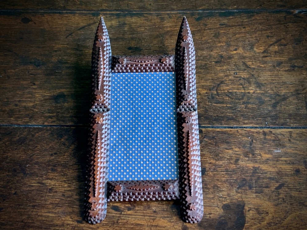 A dark brown hand-carved tramp art wooden picture frame featuring notch-carved geometric borders and pointed finials encasing a monochrome portrait photograph. View VI