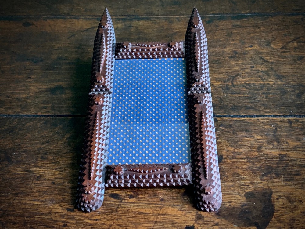 A dark brown hand-carved tramp art wooden picture frame featuring notch-carved geometric borders and pointed finials encasing a monochrome portrait photograph. View VII