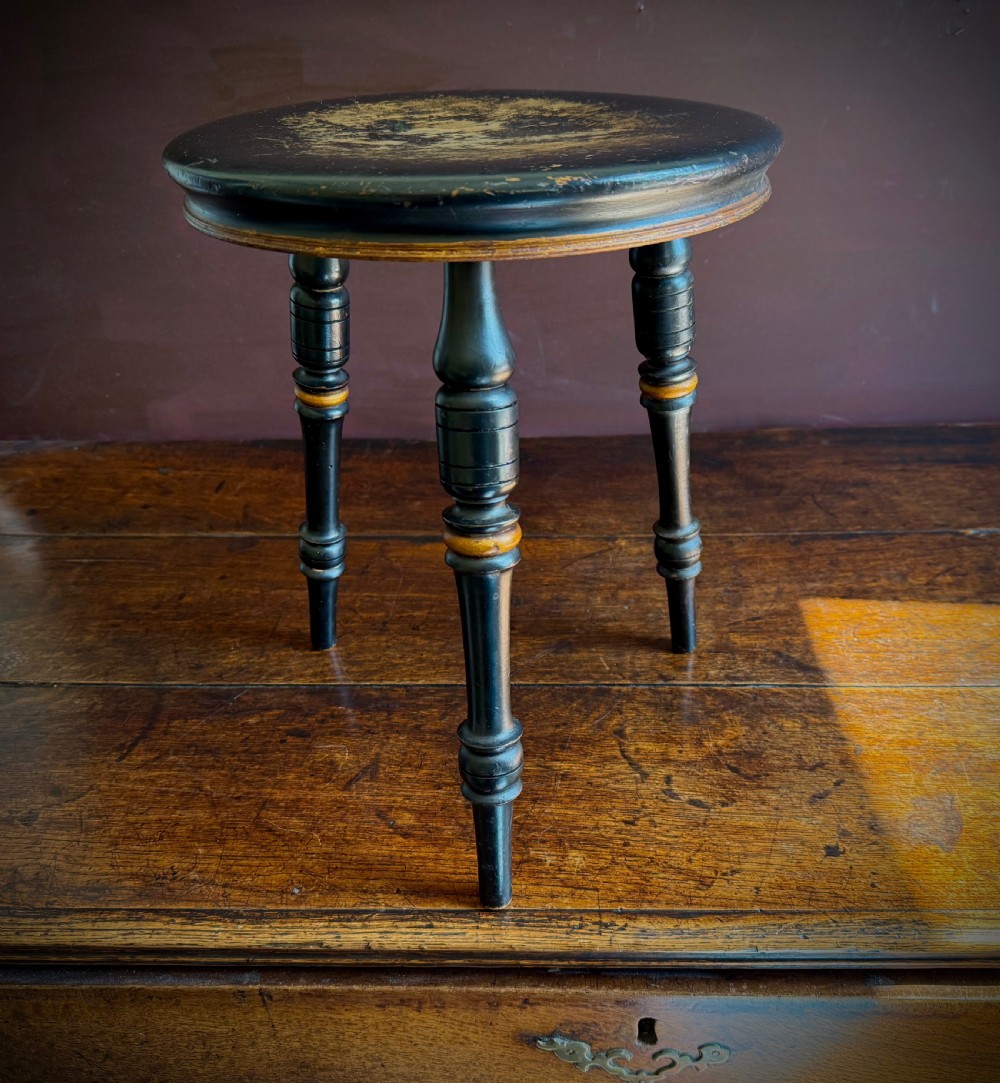 A Victorian ebonised three-legged stool featuring turned legs with gilt band highlights and a circular top showing surface wear.