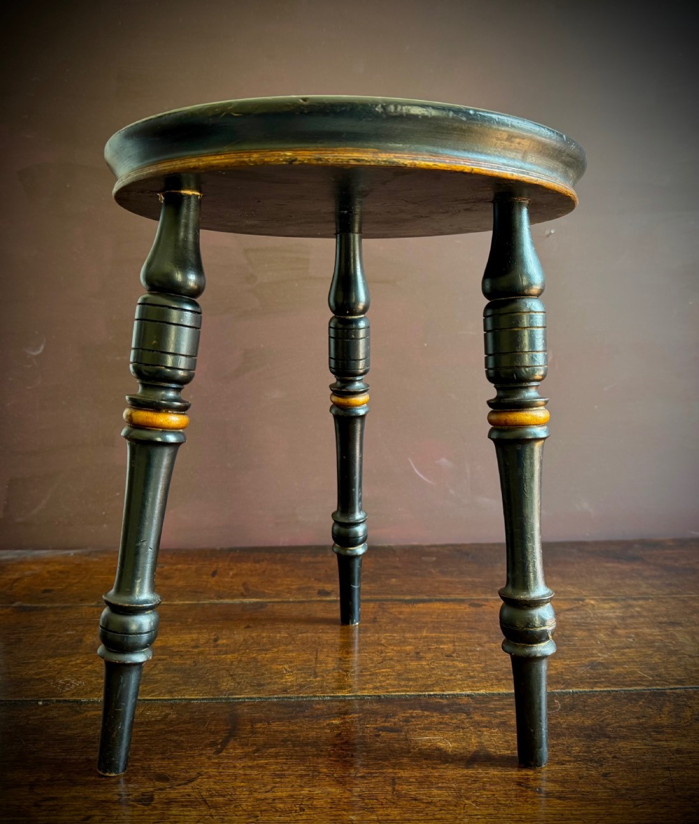 A Victorian ebonised three-legged stool featuring turned legs with gilt band highlights and a circular top showing surface wear. View II