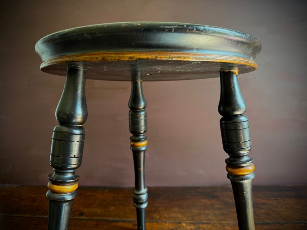 A Victorian ebonised three-legged stool featuring turned legs with gilt band highlights and a circular top showing surface wear. View IV