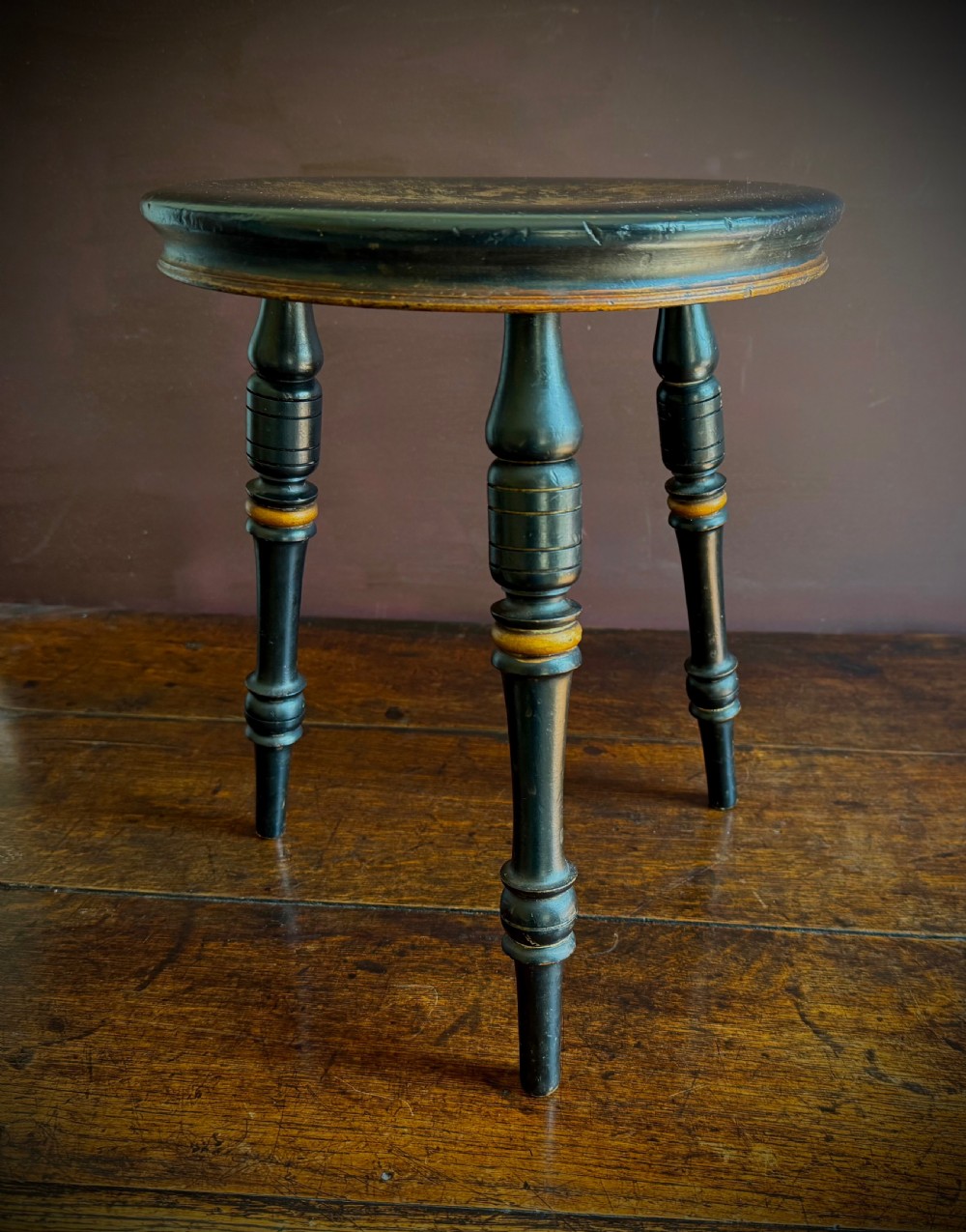 A Victorian ebonised three-legged stool featuring turned legs with gilt band highlights and a circular top showing surface wear. View VI