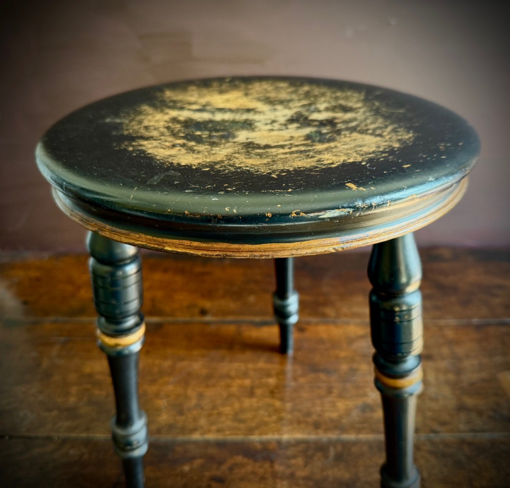 A Victorian ebonised three-legged stool featuring turned legs with gilt band highlights and a circular top showing surface wear. View IX