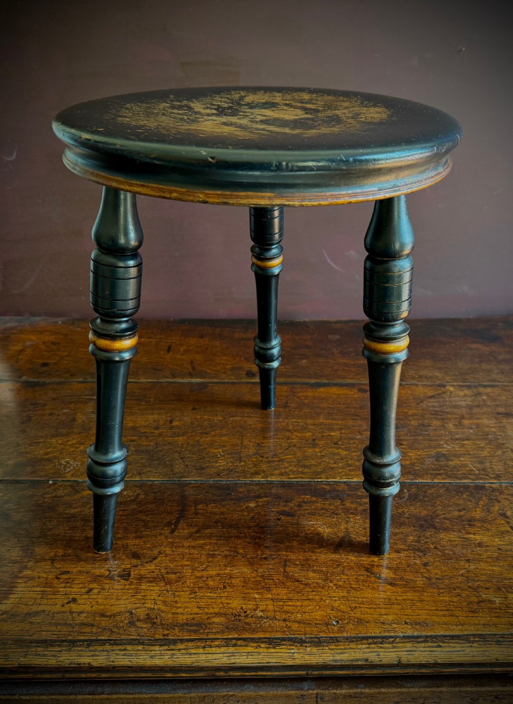 A Victorian ebonised three-legged stool featuring turned legs with gilt band highlights and a circular top showing surface wear. View X