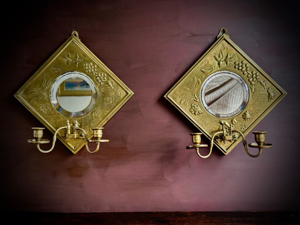 A pair of diamond-shaped embossed brass girandole wall mirrors featuring Chinoiserie scenes and twin candle branches.