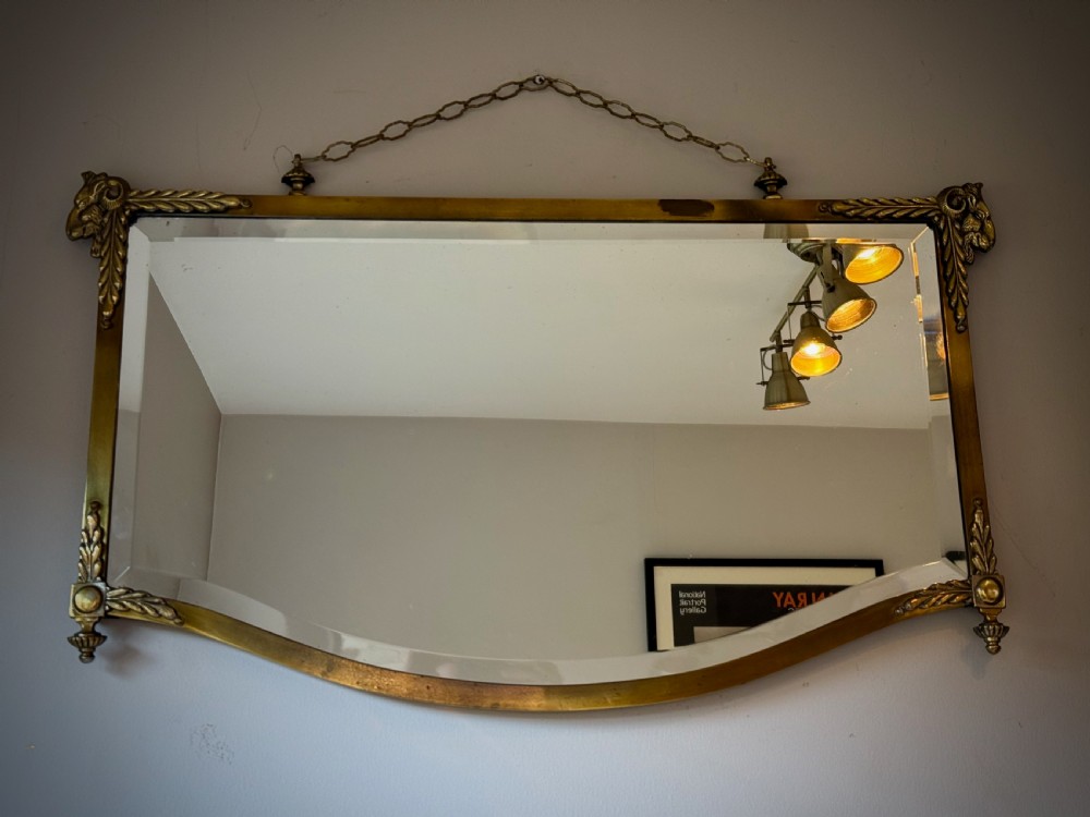 Horizontal wall mirror featuring a brass frame with sculpted rams heads on the top corners, leaf motifs, a curved lower edge, and an overhead hanging chain.
